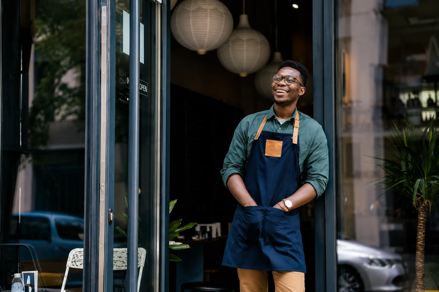 A café owner in an apron standing in the doorway of his shop.