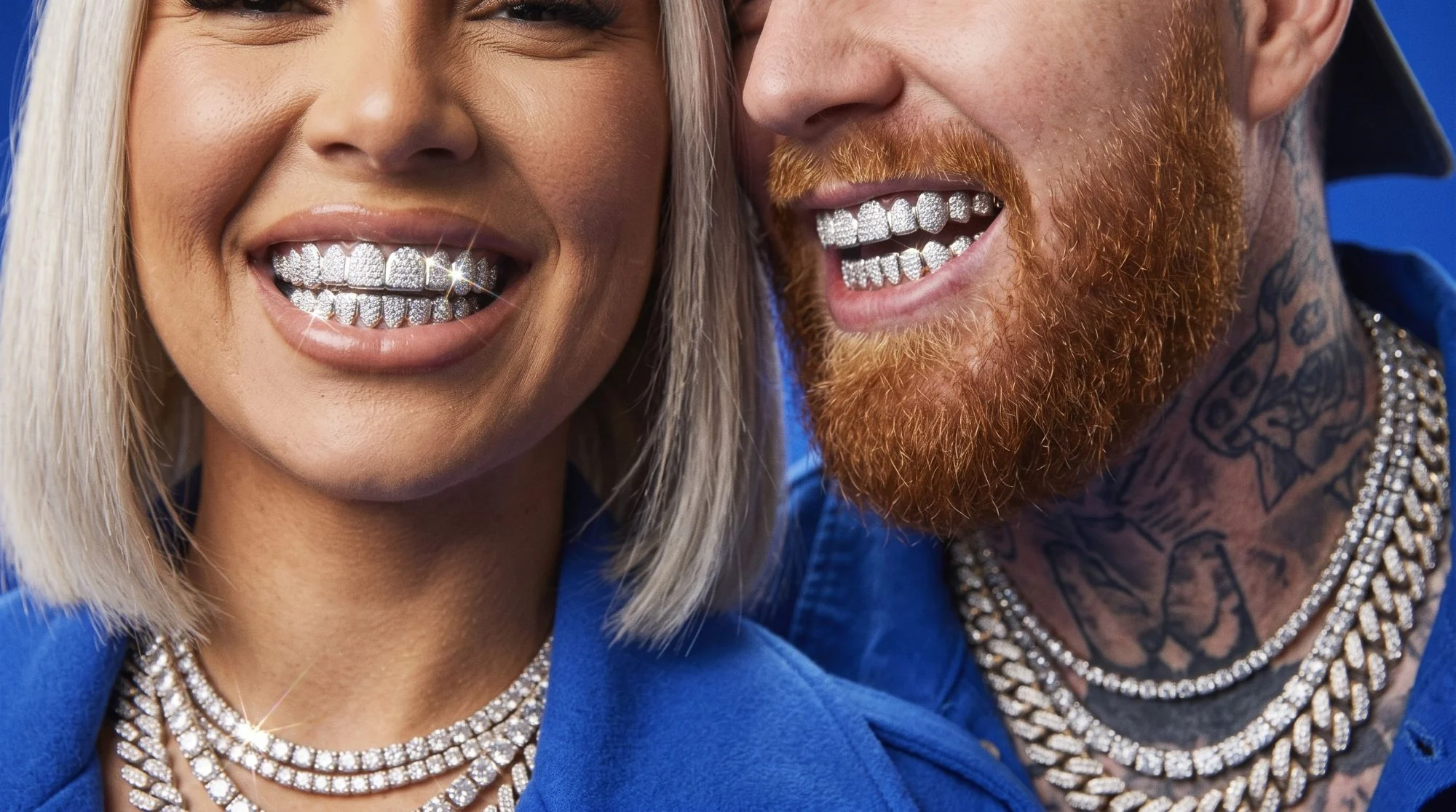 Close-up of a woman and a red-bearded man smiling in iced-out grillz and diamond chains, against cobalt blue