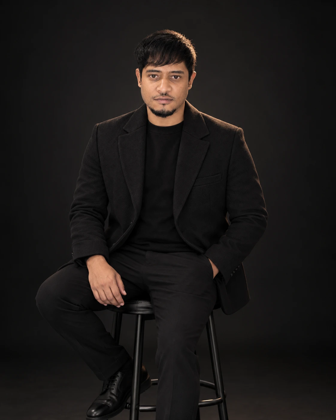 Shaheed Gqokoma seated on a stool in black against a dark backdrop