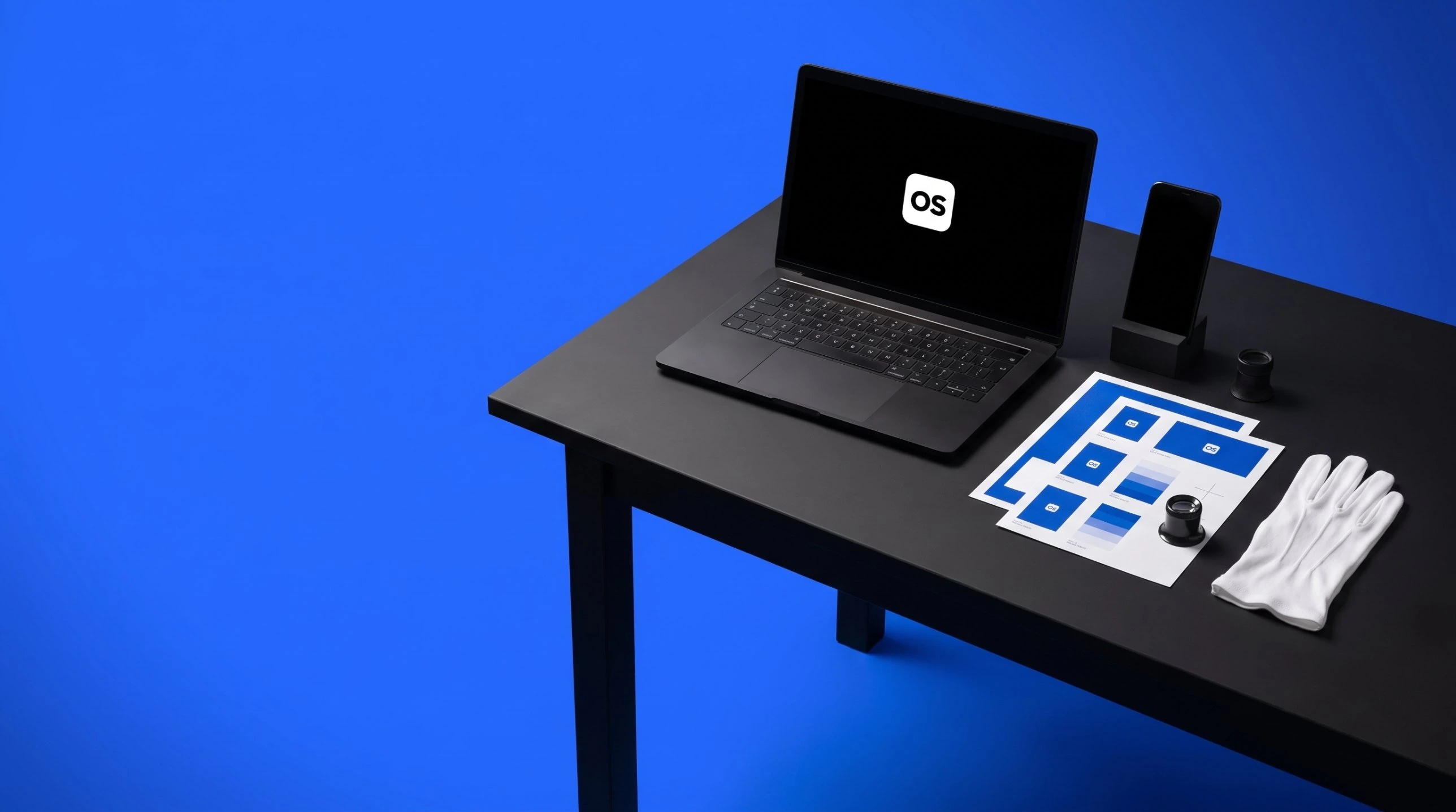 A black worktable on a cobalt floor laid out with a laptop, a phone on a stand, printed proofs, a loupe and white gloves